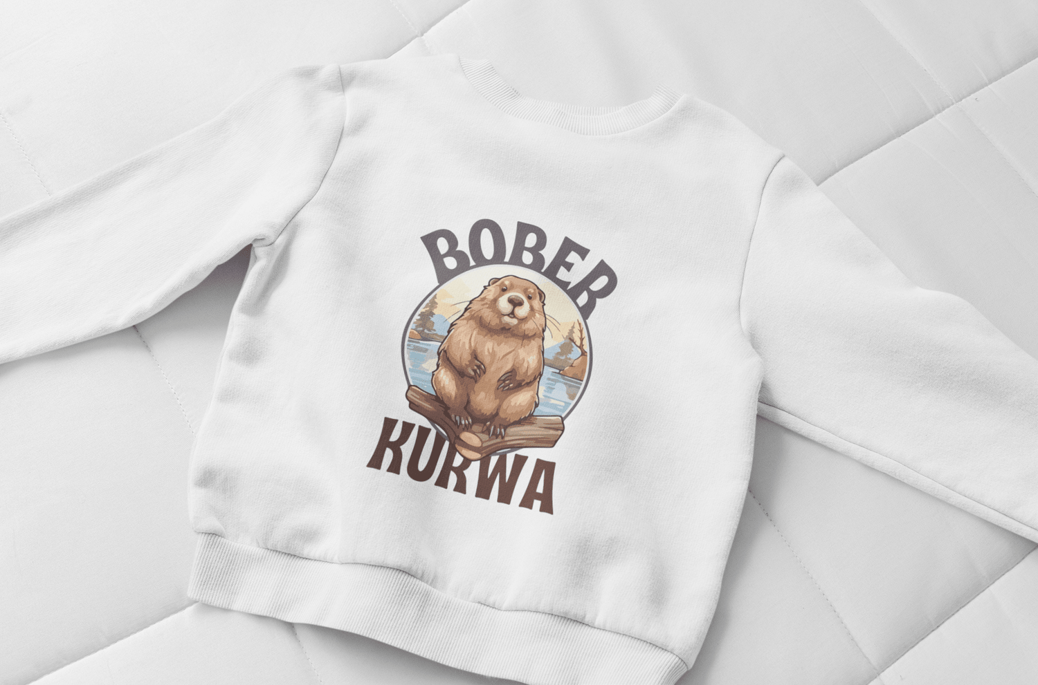 A white long-sleeve sweatshirt laid flat on a white quilted surface, featuring a central graphic of a brown beaver sitting on a log with the words 'BOBER KURWA' in dark gray and brown lettering.