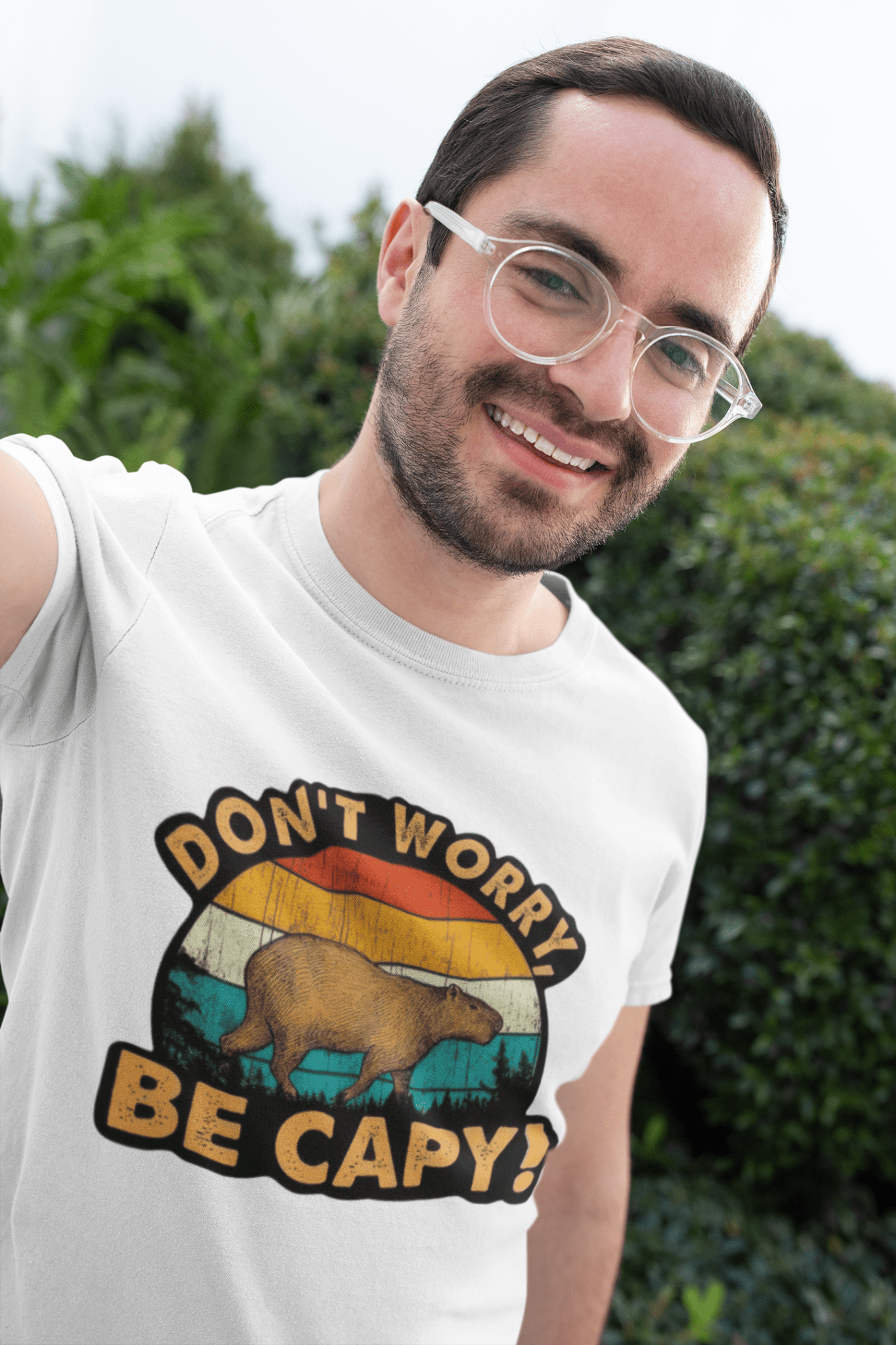 A man wearing a white graphic t-shirt with a retro-style bear design and the text 'DON'T WORRY BE CAPY!' against a blurred green outdoor background.